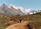Ein Pfad mit einer spazierenden Person. In Hintergrund befindet sich eine blaue Htte und Berge die an den Wolken kratzen